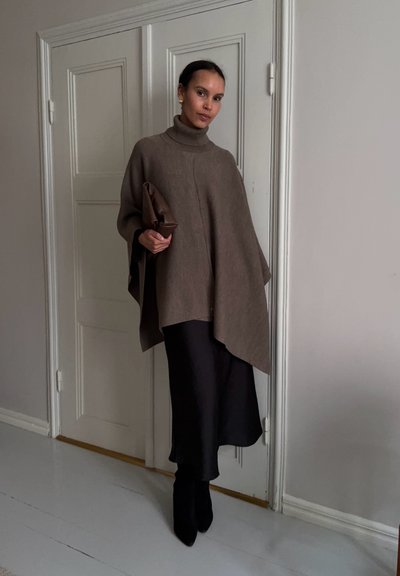 Mujer con poncho de cuello alto marrón, falda midi negra, botas negras, sosteniendo un clutch marrón, de pie en el interior junto a una puerta blanca con paneles.