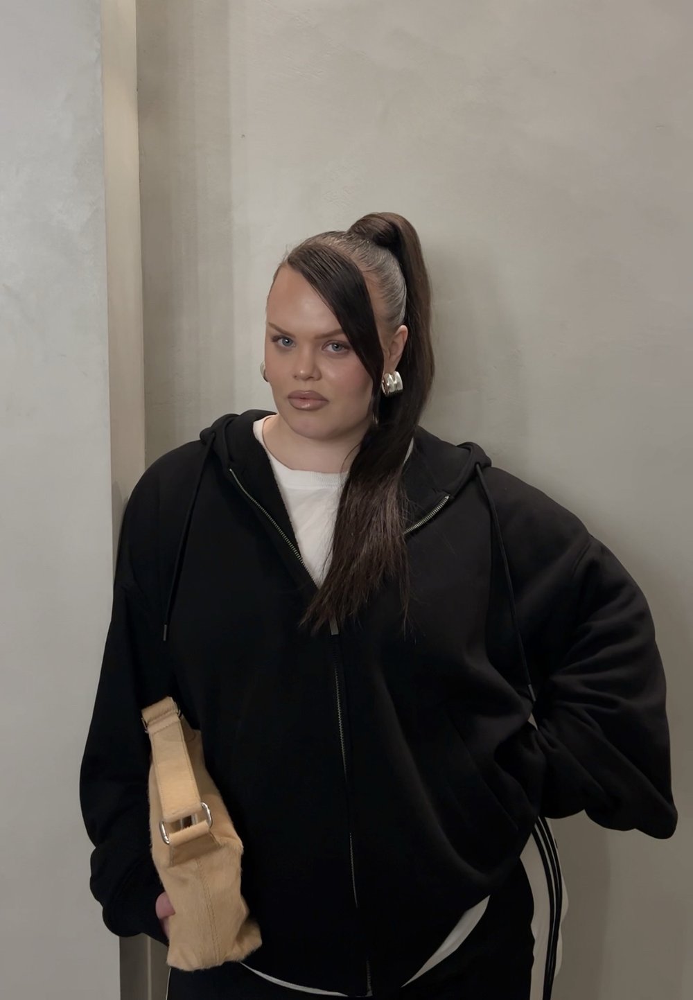 Sudadera con cremallera negra sobre una camiseta blanca, combinada con pantalones negros. Sosteniendo un bolso beige claro, con el cabello largo y liso recogido en una coleta alta.