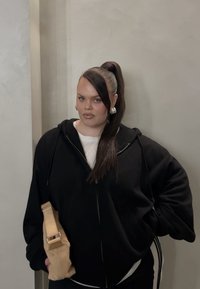 Sudadera con cremallera negra sobre una camiseta blanca, combinada con pantalones negros. Sosteniendo un bolso beige claro, con el cabello largo y liso recogido en una coleta alta.