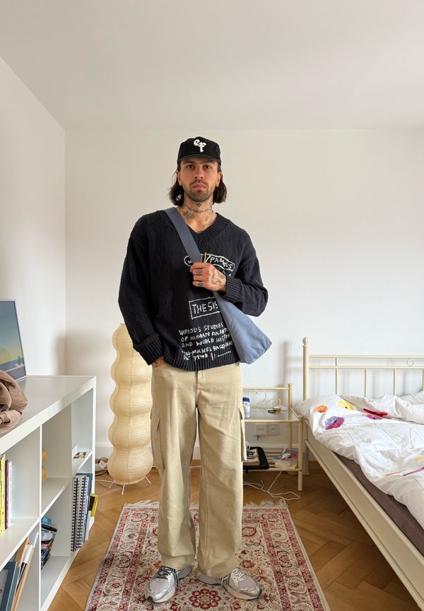 Homme portant une casquette noire, un pull noir avec un texte blanc, un pantalon beige et des baskets argentées se tient dans une chambre avec lit, lampe, bibliothèque et tapis.