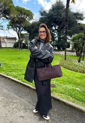 Chaqueta de cuero negra oversized, suéter de cuello alto gris, pantalones de rayas verticales, bolso de punto marrón, zapatillas casuales en blanco y negro, fondo de parque verde.