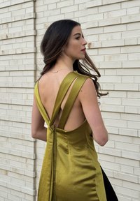 Jeune femme aux longs cheveux bruns portant une robe en satin vert olive sans dos, debout de côté contre un mur en briques clair.