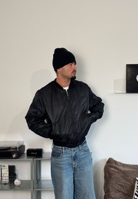 Must bomberjakk lukuga, kantud valge särgi kohal, koos helesiniste lõdvestunud teksade ja musta mütsiga.