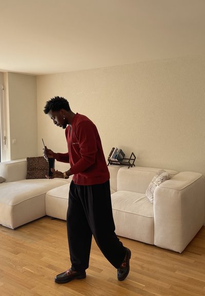 Hombre con suéter rojo y pantalones negros sosteniendo un portátil negro, de pie en una sala con sofá beige y suelo de madera.