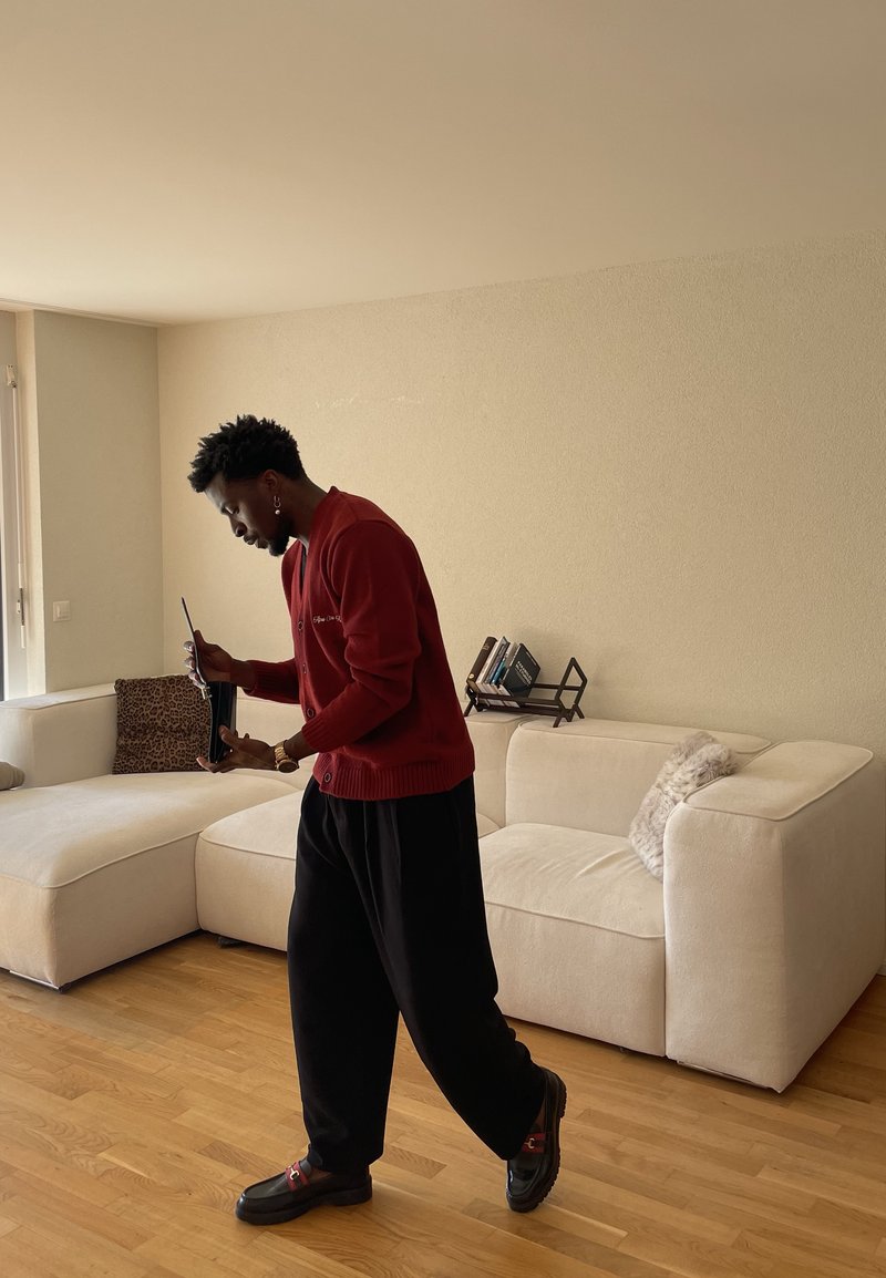 Homme en pull rouge et pantalon noir tenant un ordinateur portable noir, debout dans un salon avec un canapé beige et un sol en bois.