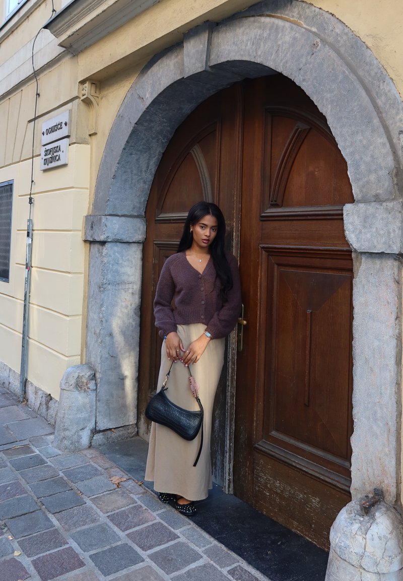 Καφέ πλεχτό κιμονό, μπεζ μακρύ φούστα, μαύρη τσάντα χειρός με υφασμάτινο σχέδιο και πιστωμένες μπαλαρίνες, ποζάρει μπροστά από μια ξύλινη πόρτα με πέτρινη καμάρα.