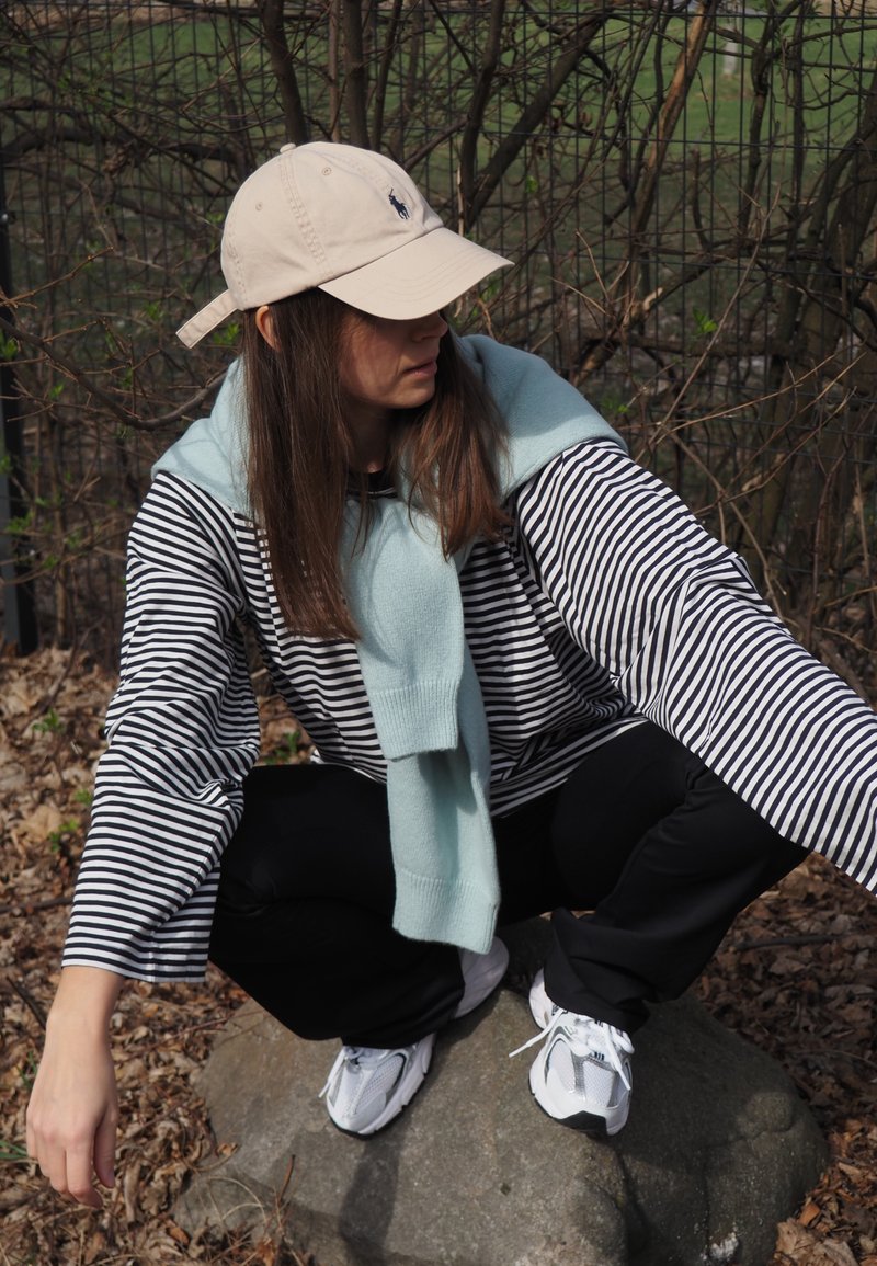 Jeune personne en chemise à rayures et casquette blanche accroupie sur un rocher en plein air, avec un pull bleu clair posé sur les épaules.