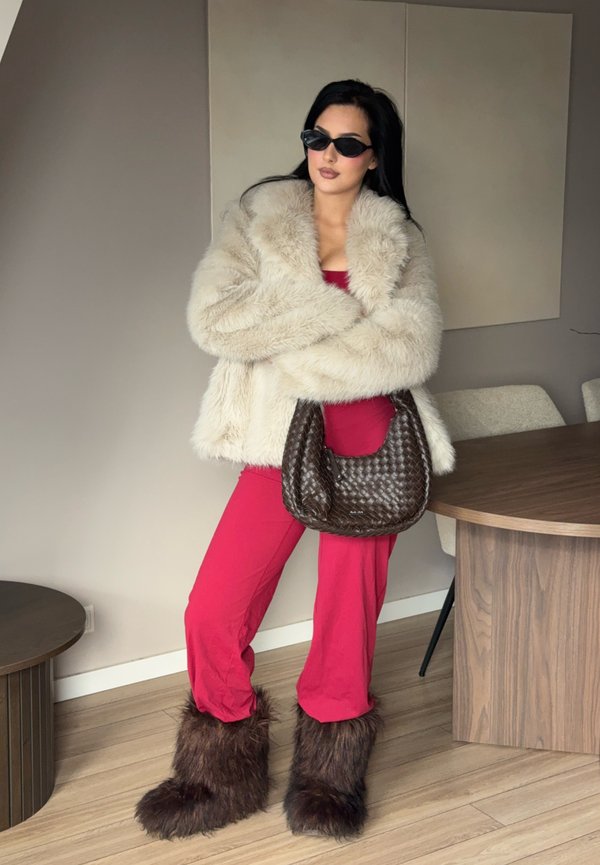 Žena v béžovom umelom kožuchu, červenom oblečení, hnedých kožušinových čižmách, tmavých slnečných okuliaroch a držiaca veľkú hnedú pletenú kabelku v interiéri.