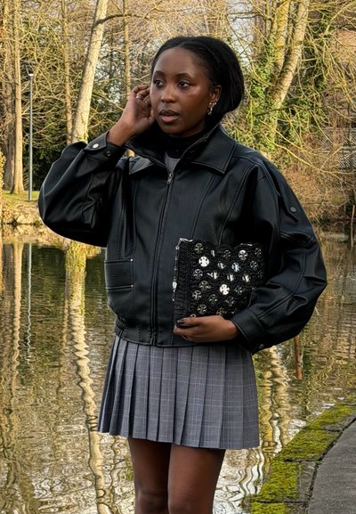 Mujer con chaqueta de cuero negra y falda plisada gris, sosteniendo un bolso de mano negro adornado, de pie junto a un estanque con árboles desnudos al fondo.