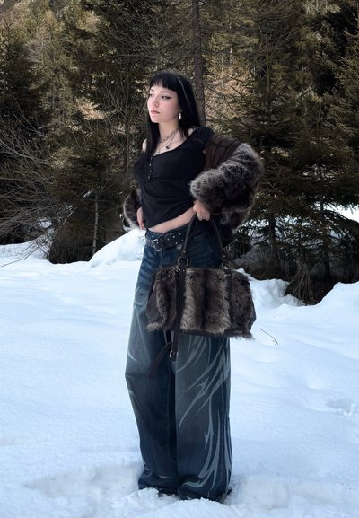 Joven mujer en un bosque nevado viste una camiseta negra, jeans de pierna ancha con estampado, chaqueta de piel en los brazos y lleva un bolso grande de piel.