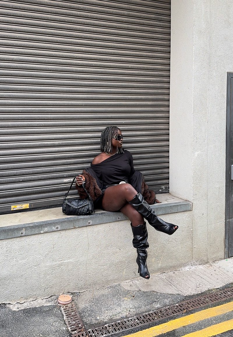 Woman in black off-shoulder dress, knee-high boots, and sunglasses sitting on concrete ledge by metal shutter, holding studded black handbag.