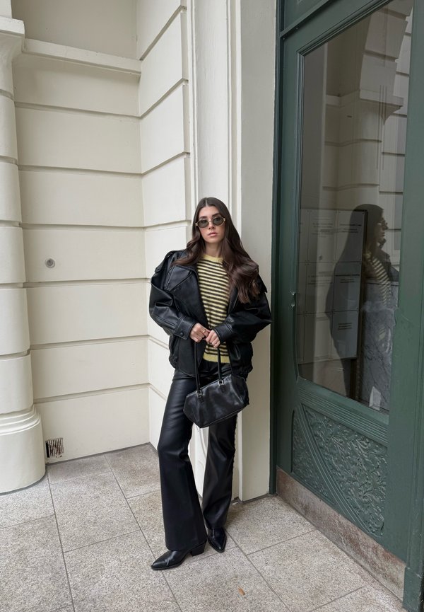 Ung kvinde iført sort læderjakke, sorte læderbukser, stribet sweater, solbriller og med en sort håndtaske, lænende sig op ad en hvid væg.