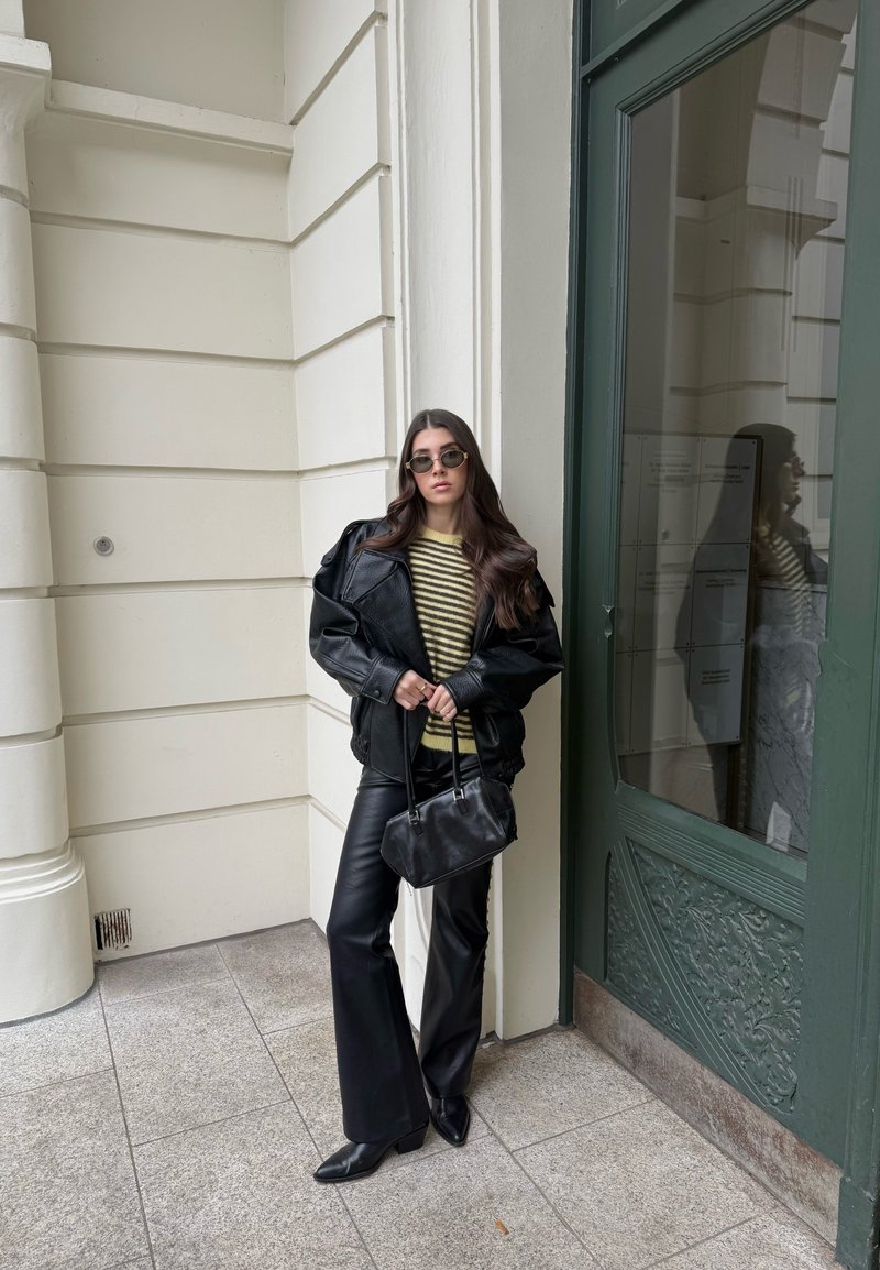 Giovane donna con giacca di pelle nera, pantaloni di pelle neri, maglione a righe, occhiali da sole e borsa nera in mano, appoggiata a un muro bianco.
