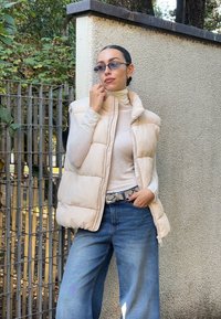 Bežinė pūkinė liemenė virš šviesios golfo tipo palaidinės, derinama su plačiomis mėlynomis džinsinėmis kelnėmis. Faktūrinis sienos ir tvoros apšvietimas pabrėžia lauko aplinką.