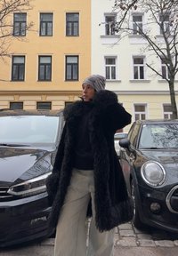 Long manteau en fausse fourrure noire avec une finition texturée et un large col, associé à un pull foncé et un pantalon ample clair. Bonnet gris à motifs.
