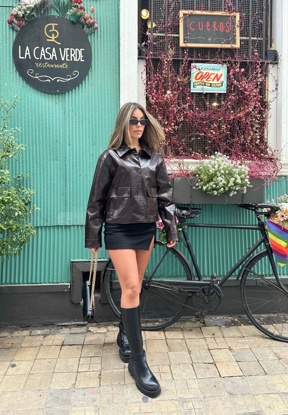 Frau in schwarzer Lederjacke, Minikleid und Stiefeln, die an einem türkisfarbenen Gebäude mit einem Fahrrad und Blumendekorationen vorbeigeht.