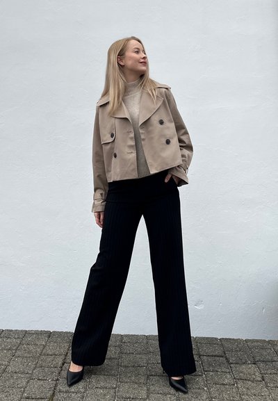 Mujer rubia vestida con chaqueta beige corta, suéter ligero, pantalones de pierna ancha negros y tacones de punta negros, de pie contra una pared blanca.