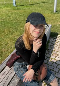 Svart slitna keps, långt blont hår, svart långärmad tröja, denimshorts, håller en basketboll, silverringar, sitter på en bänk av trä.