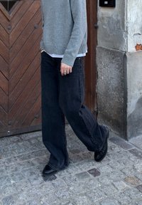 Pull en maille gris, jeans larges noirs et chaussures noires. Debout sur des pavés, avec une porte en bois et un mur en pierre texturé en arrière-plan.