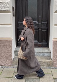 Lange, bruine, gestructureerde wollen jas met grote zakken; draagt een bruine handtas met patroon. Zwarte broek met witte accenten op de schoenen; stedelijke omgeving.