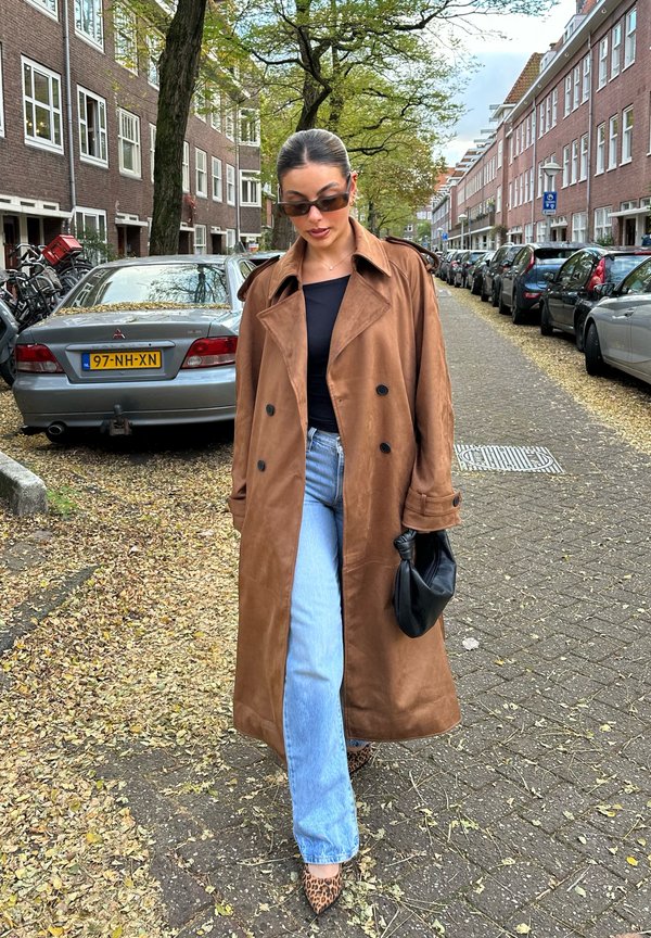 Trench coat de camurça castanho com botões grandes, combinado com jeans de perna larga azul claro e sapatos de salto de padrão leopardo. Bolsa preta na mão.