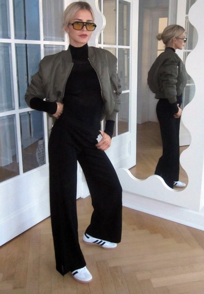 Chaqueta bomber verde oliva sobre un cuello de tortuga negro, combinado con pantalones de pierna ancha negros y zapatillas blancas con rayas negras. Fondo de espejo de cuerpo entero.