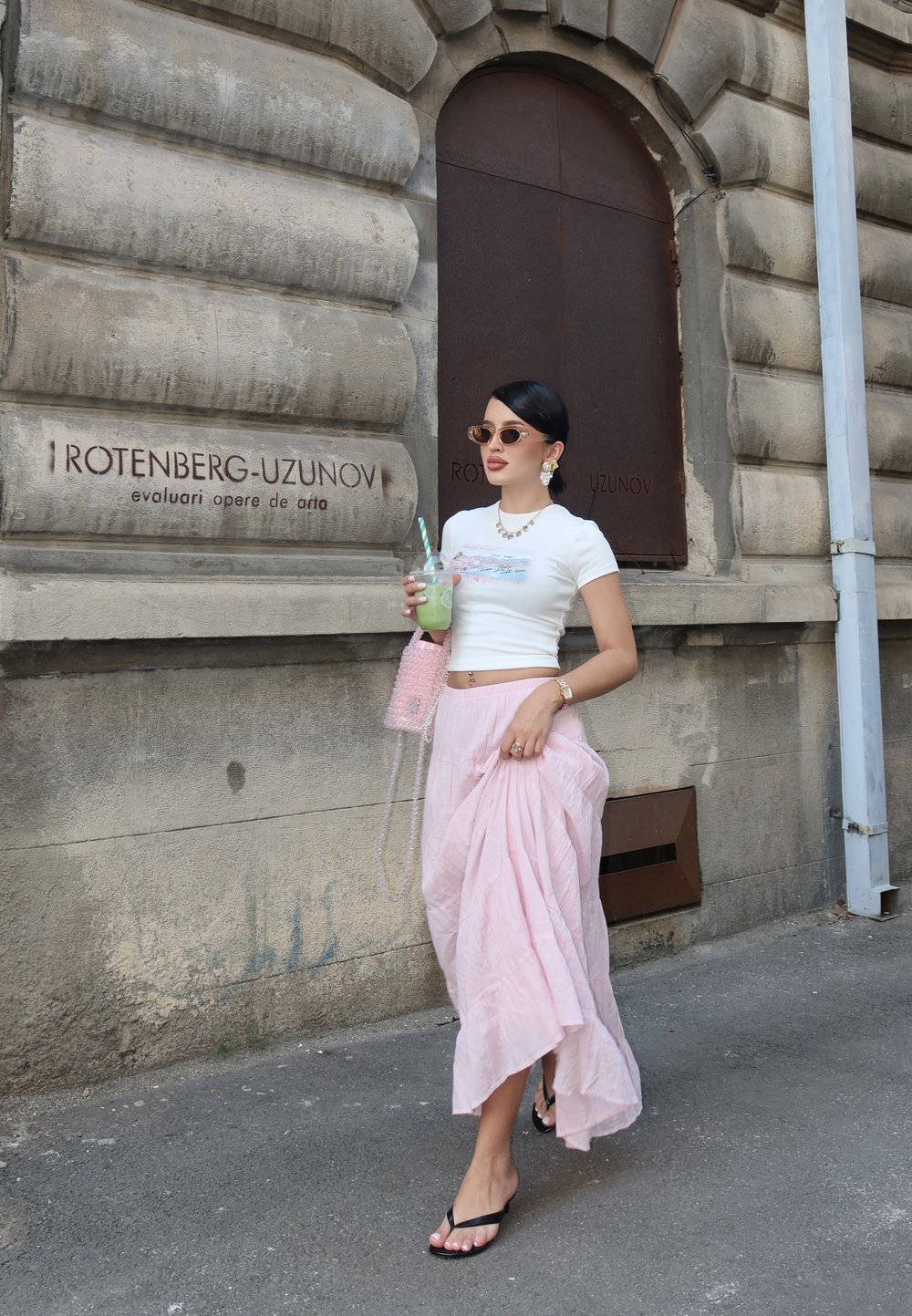 Világos rózsaszín, laza maxi szoknya fehér, crop top-pal párosítva. Kiegészítők között oversized napszemüveg, fülbevalók és egy rózsaszín gyöngyös táska szerepel.