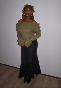Sweater verde-oliva sem ombros com detalhes em franjas, combinado com uma saia longa preta e botas de tornozelo pretas. Acessórios dourados visíveis.