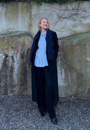 Mujer con un abrigo largo negro, blusa azul claro y pantalones negros, de pie sobre grava frente a una pared de roca cubierta de musgo, sonriendo y mirando hacia la derecha.