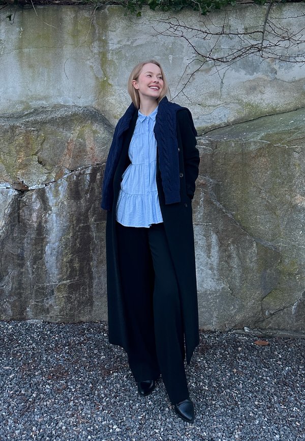 Frau in langem schwarzem Mantel, hellblauer Bluse und schwarzer Hose steht auf Kies vor einer moosbedeckten Felswand, lächelt und schaut nach rechts.