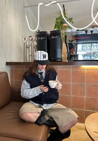 Personne portant une chemise rayée, un gilet et une casquette, assise sur un canapé marron tenant une tasse de café dans un café moderne avec des machines à café en arrière-plan.