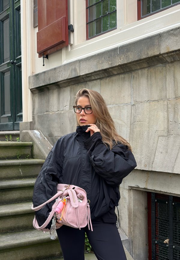 Mujer con chaqueta negra y gafas sosteniendo un bolso rosa, de pie cerca de unos escalones de piedra y un edificio con grandes ventanas.