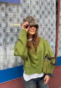 Sweatshirt verde de tamanhos grandes sobre uma camisa branca; jeans cinzentos; chapéu felpudo de padrão leopardo; pequena bolsa de camurça verde com ferragens prateadas.