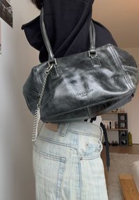 Personne portant un jean bleu clair et un haut noir, avec un sac à main en cuir noir Liebeskind Berlin orné d'un charme peigne argenté, posé sur l'épaule.