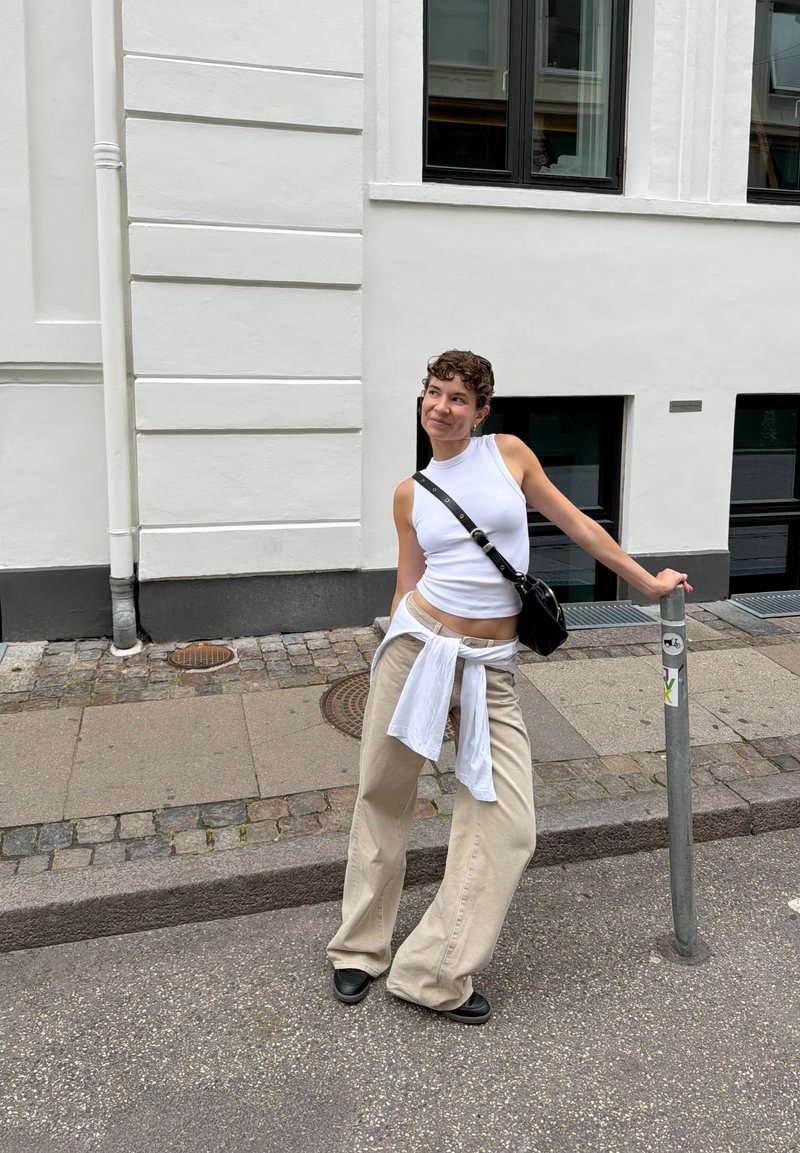 Junge Frau in weissem ärmellosem Oberteil und beigen Hosen lehnt an einem Metallpfosten auf einer Stadtstrasse mit weissem Gebäude und Fenstern im Hintergrund.
