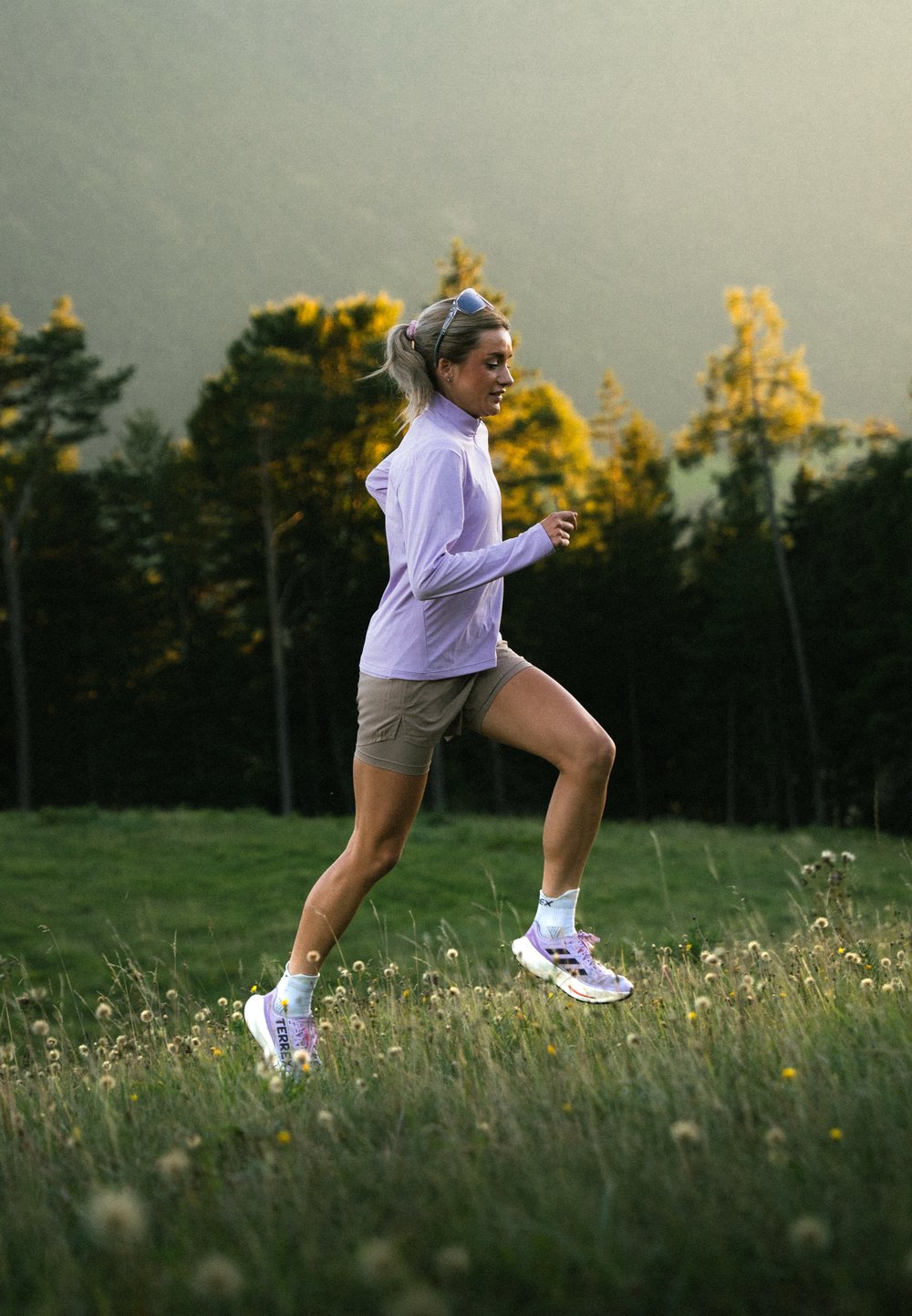 Lys lilla langærmet skjorte, beige shorts, løbende på græs. Hvide sko med lilla og orange accenter. Baggrunden har træer og sollys.