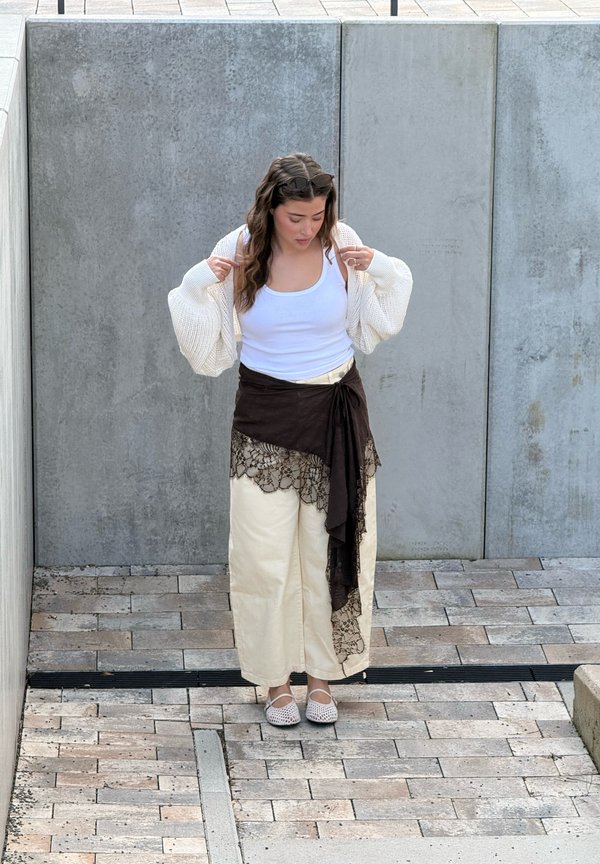 Mladá žena si upravuje biely kardigán cez biele tielko, má na sebe krémové nohavice so širokými nohavicami a čierny čipkovaný šál, stojí na kamennej dlažbe.