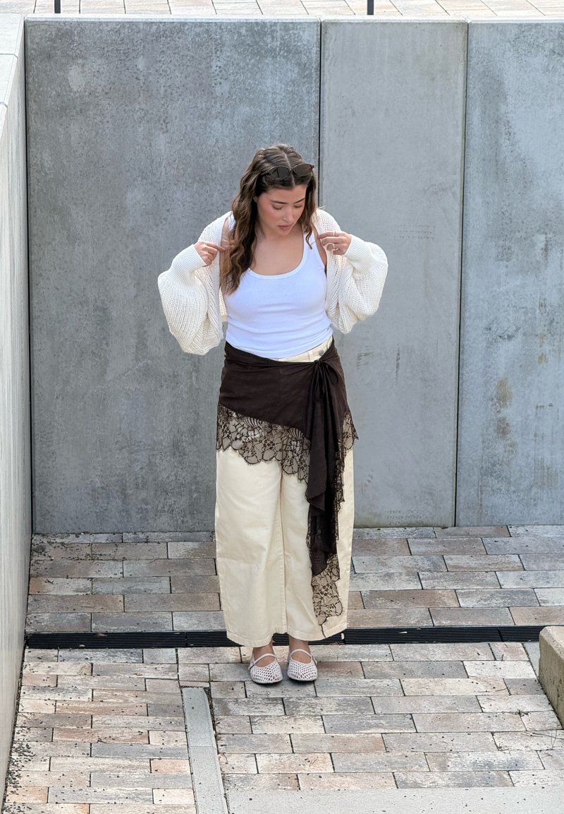 Νεαρή γυναίκα προσαρμόζει λευκή ζακέτα πάνω από λευκή φανέλα, φοράει κρεμ παντελόνια με φαρδιά μπατζάκια και μαύρο δαντελένιο φουλάρι, στέκεται σε πλακόστρωτο.