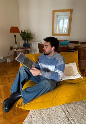 Una persona con un suéter azul y blanco está sentada en un sofá amarillo mostaza, sosteniendo una revista. El sofá tiene una tela texturizada y cojines suaves.