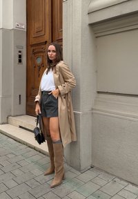 Manteau beige sur un top blanc, short gris et bottes en cuir marron jusqu'aux genoux. Sac noir avec des ferrures dorées. Debout près d'une porte en bois.