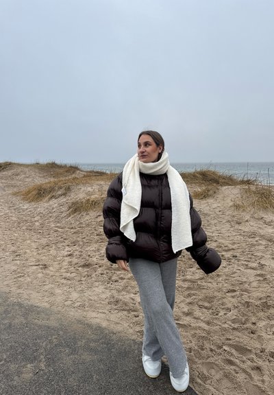 Chaquetón marrón esponjoso, bufanda de canalé color crema, pantalones grises y zapatos blancos; de pie sobre arena con una playa y el océano al fondo.