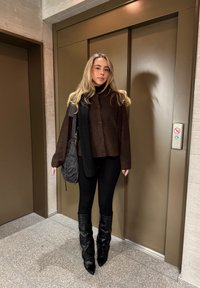 Fekete térdig érő bőr csizmák, szűk fekete nadrág, sötétbarna texturált, bő szabású, gombos ing, fekete sállal; egy lift közelében áll.