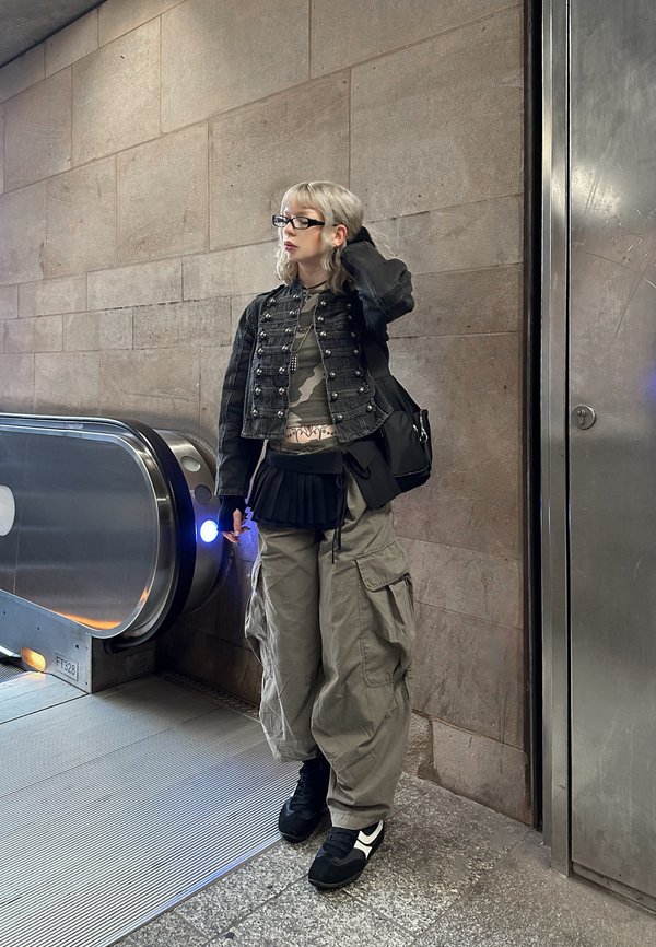Une personne portant des lunettes et des vêtements streetwear superposés se tient à côté d’un escalator dans une station de transport aux murs de pierre.