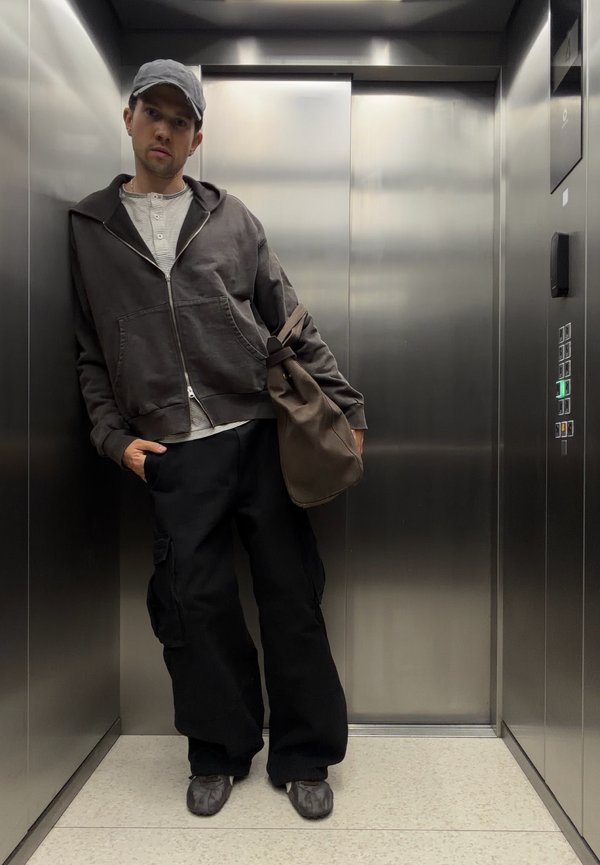 Hombre con gorra, sudadera oscura con capucha y pantalones cargo negros, llevando una bolsa marrón, de pie dentro de un ascensor de acero inoxidable.