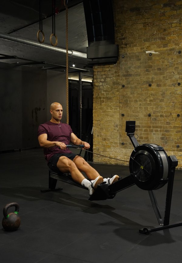 Man rowing on an indoor rowing machine in a gym with a brick wall, gymnastic rings, and a kettlebell nearby.
