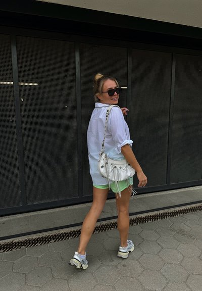 Camisa blanca de botones, pantalones cortos verdes, bolso plateado con flecos y zapatillas grises. Unas gafas de sol negras completan el look casual.