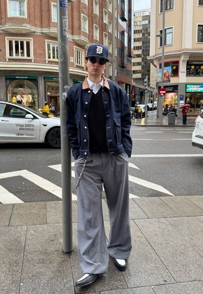 Chaqueta navy con cuello beige sobre un suéter negro, combinada con pantalones anchos de rayas grises y zapatos con detalles plateados, apoyado contra un poste de la calle.