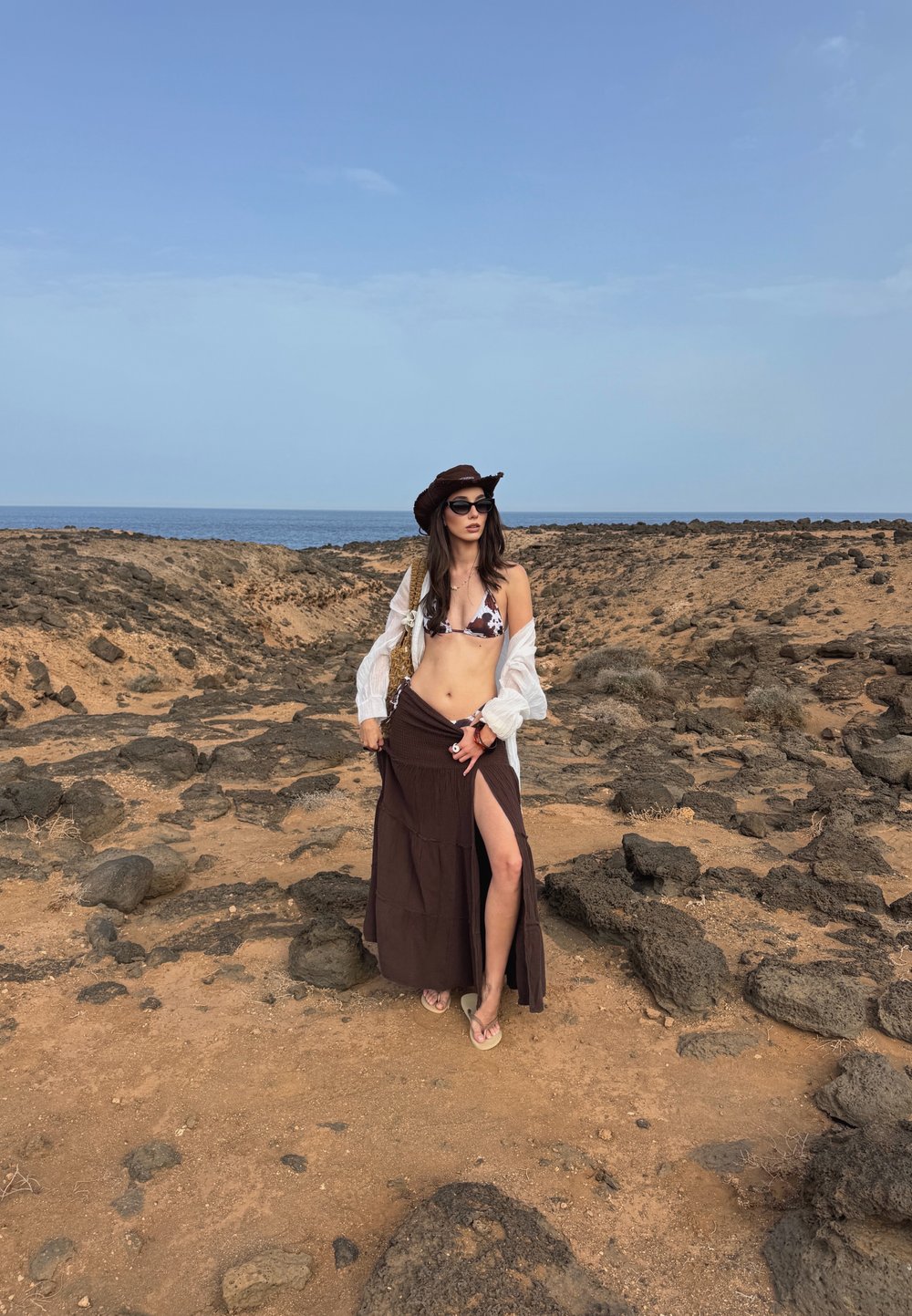 Brun maxi skjørt med høyt splitt, hvit gjennomsiktig skjorte og dyreprint-bikini topp. Tilbehør inkluderer en brun hatt og solbriller.