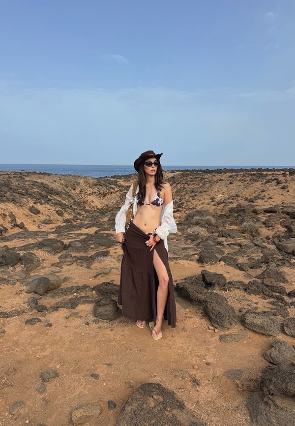 Saias maxi castanha com uma fenda até à coxa, camisa branca transparente e parte de cima de biquíni com padrão animal. Os acessórios incluem um chapéu castanho e óculos de sol.
