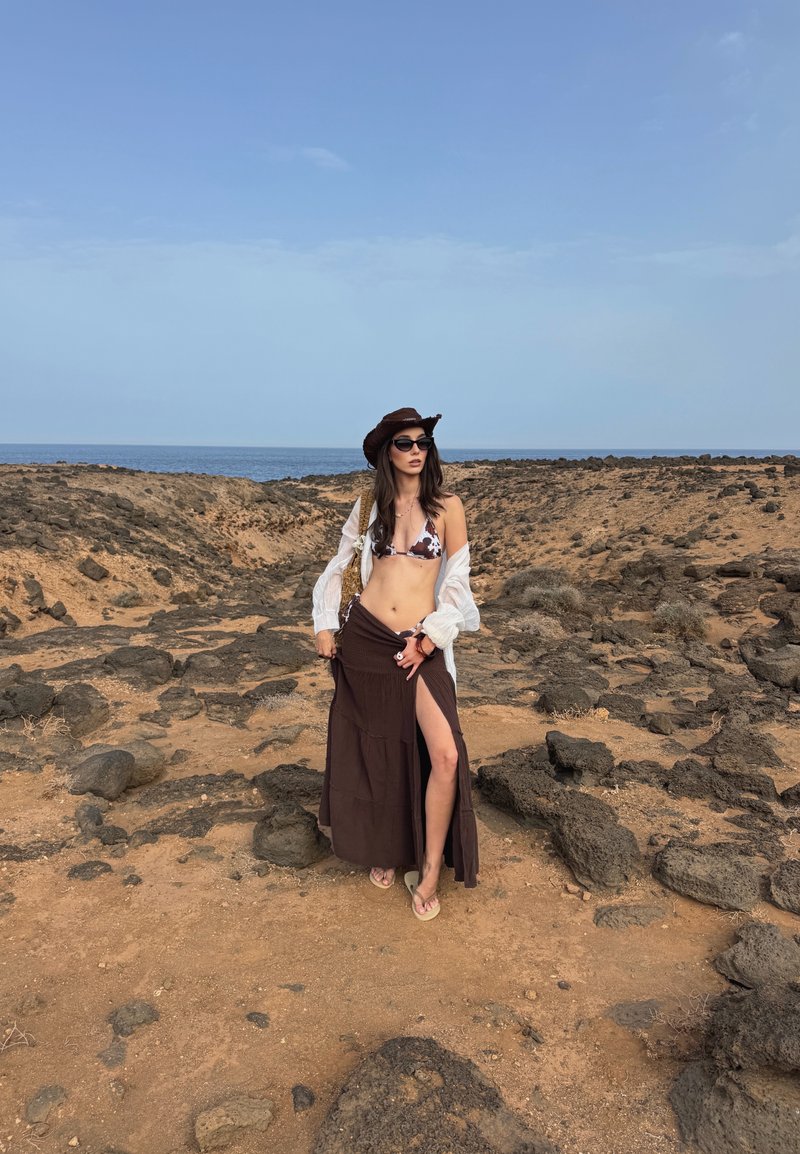 Brun maxikjol med en hög slits till låret, vit genomskinlig skjorta och bikiniöverdel med djurmönster. Tillbehör inkluderar en brun hatt och solglasögon.